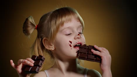 happy smiling little child girl kid eating milk chocolate bar dessert isolated on dark background