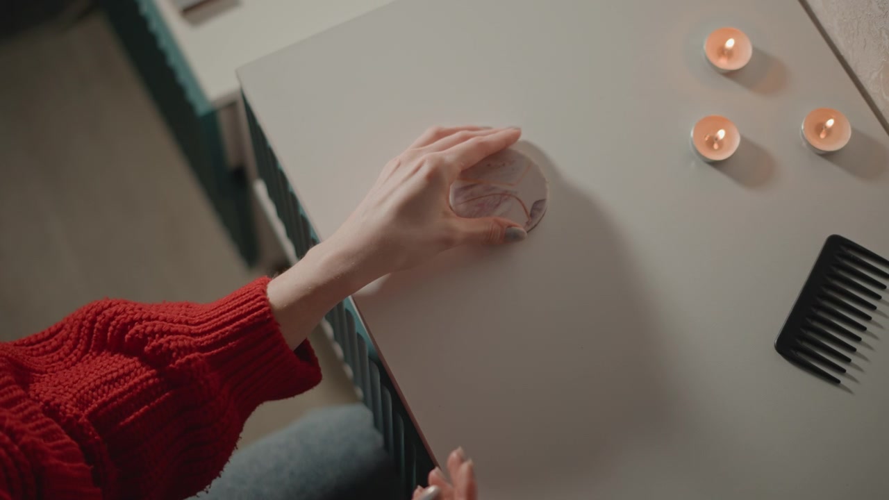 Vista dall'alto di mani adulte con un maglione di maglia rosso che aprono, chiudono e posizionano uno specchietto compatto rotondo con custodia argentata su un tavolo chiaro, accanto a candele tealight accese e un pettine, creando una scena di bellezza accogliente.