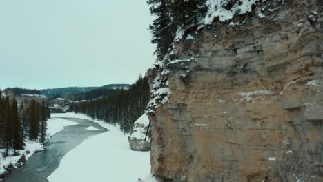 Drone-shot-rising-up-the-side-of-a-snowy-cliff-face-with-a-frigid-creek-below-with-Canadian-goose-in-the-water