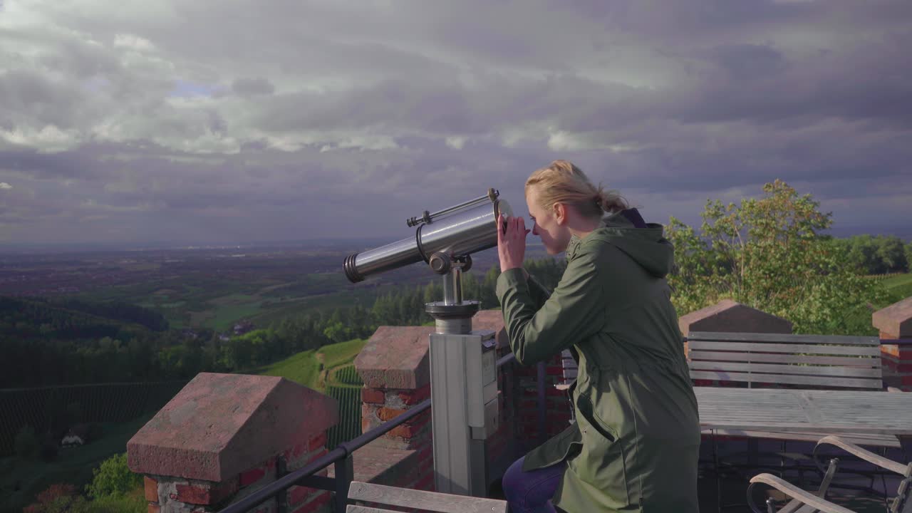 una chica rubia caminando hacia un telescopio desde un patio que pertenece a un castillo en la cima de una montaña