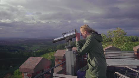 a blonde girl walking towards a telescope from a patio which belongs to a castle on top of a mountain