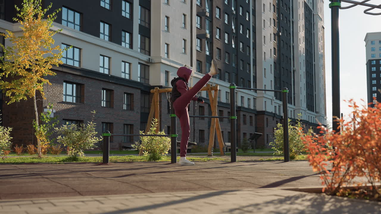 Jogger holding pull up bar while stretching her leg behind in urban outdoor workout under bright sun with residential buildings, and autumn shrubs around paved court showing warm up routine