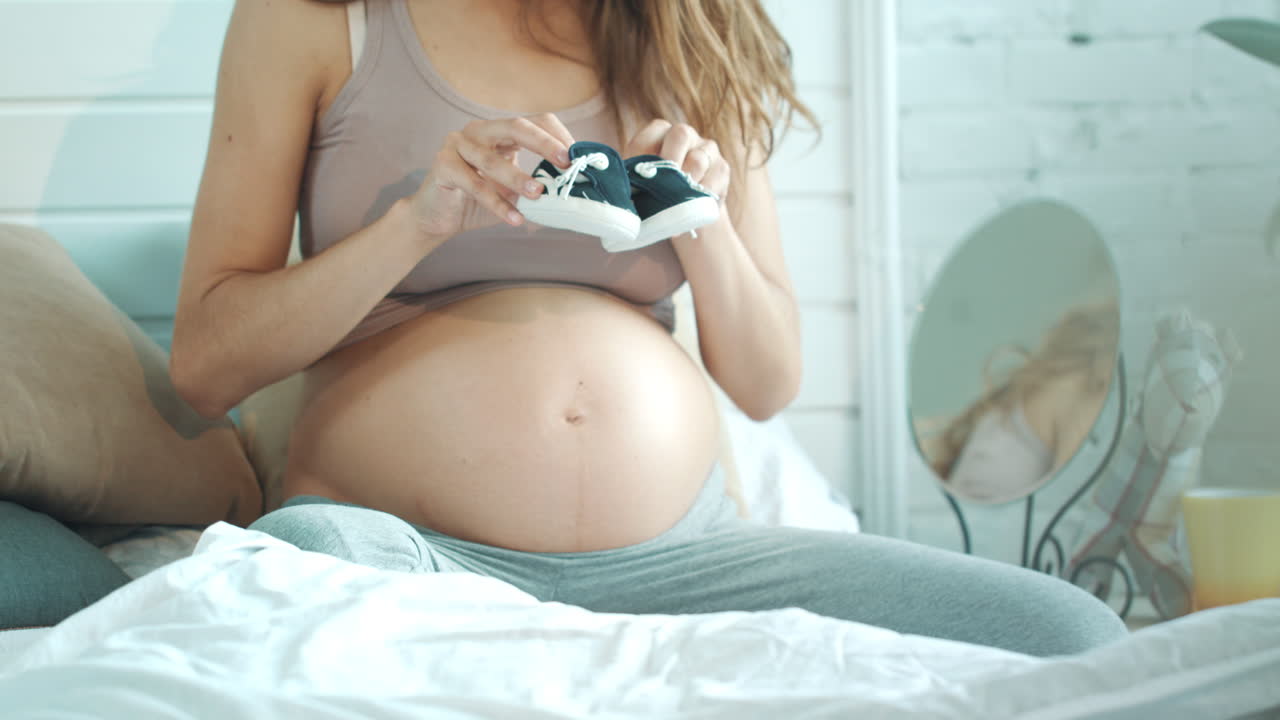 mujer embarazada no reconocida jugando con zapatos de bebé en la cama.