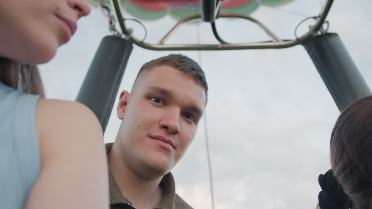 Close up two passengers looking over edge of hot air balloon basket beneath colorful envelope panels, soft evening light casting gentle shadows as balloon floats above green landscape