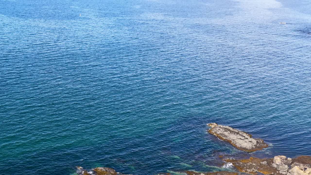 Drone ascends over Scottish lighthouse, rocky coast, tents, and blue ocean under bright daylight