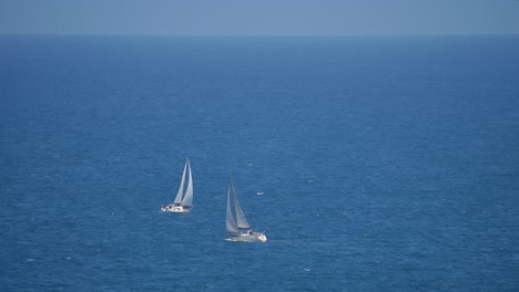 two white sailboats sailing on open calm blue sea with visible waves and distant horizon