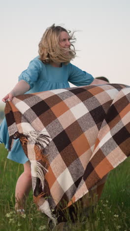 a woman in a blue dress tries to spread a large scarf on the grass as the wind catches it. her two young sons, wearing white shirts, stand nearby, gazing at the lake in the distance