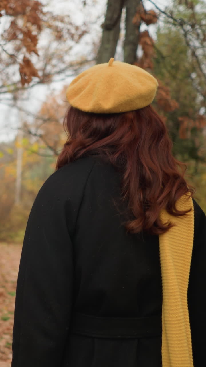 Dame i svart kåpe, gul beret og gult skjerf berører tørt løvverk, og ser seg spent rundt i en levende høstskog dekket av falne blader.