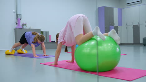 kids actively engaged in a workout session seen through a mirror, one child balances on a green exercise ball, another crawls on a purple mat using yellow balance pods