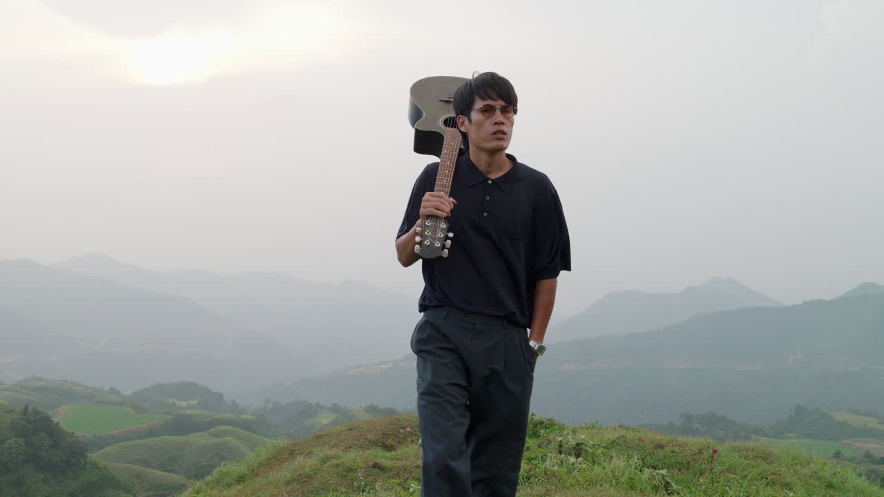 고급스러운 성숙한 아시아 남성이 열대  ⁇ 에서 해가 지는 동안 어깨에 기타를 들고  ⁇ 고 있습니다.