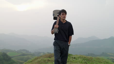 classy mature asian male walking carrying his guitar on shoulders at sunset in tropical fields