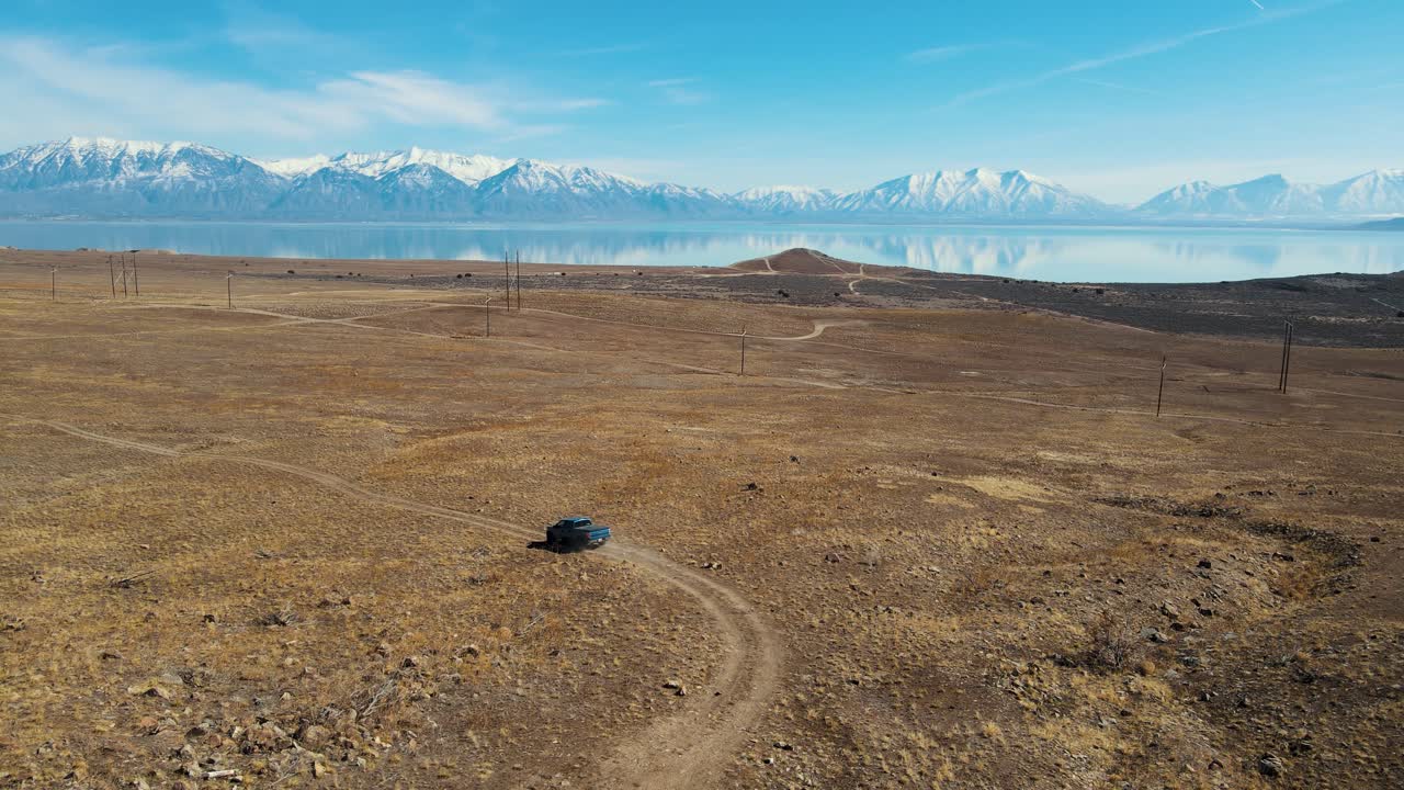 un camión azul conduciendo por un sendero todoterreno con un enorme lago y montañas nevadas a lo lejos