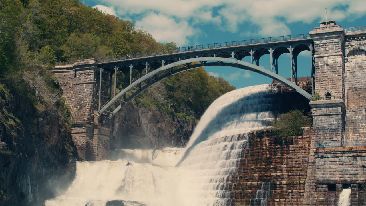 wide shot of bridge at the top of new croton dam with stepped spillway, trees and blue sky in the background. Slow motion 40fps. static
