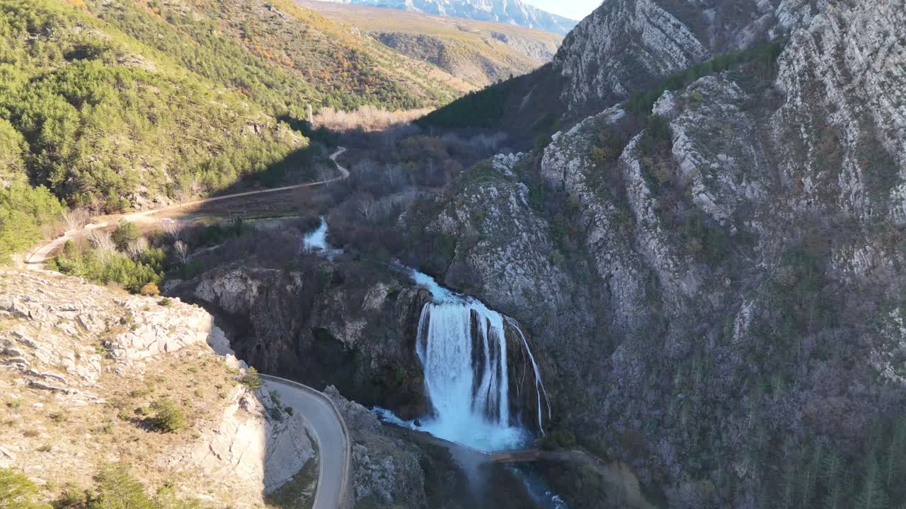Drone flying backward from Krčić Waterfall, revealing an ever-widening panorama of the Krka River, Napoleon road, curving gorge, and massive karst mountains near Knin, Croatia