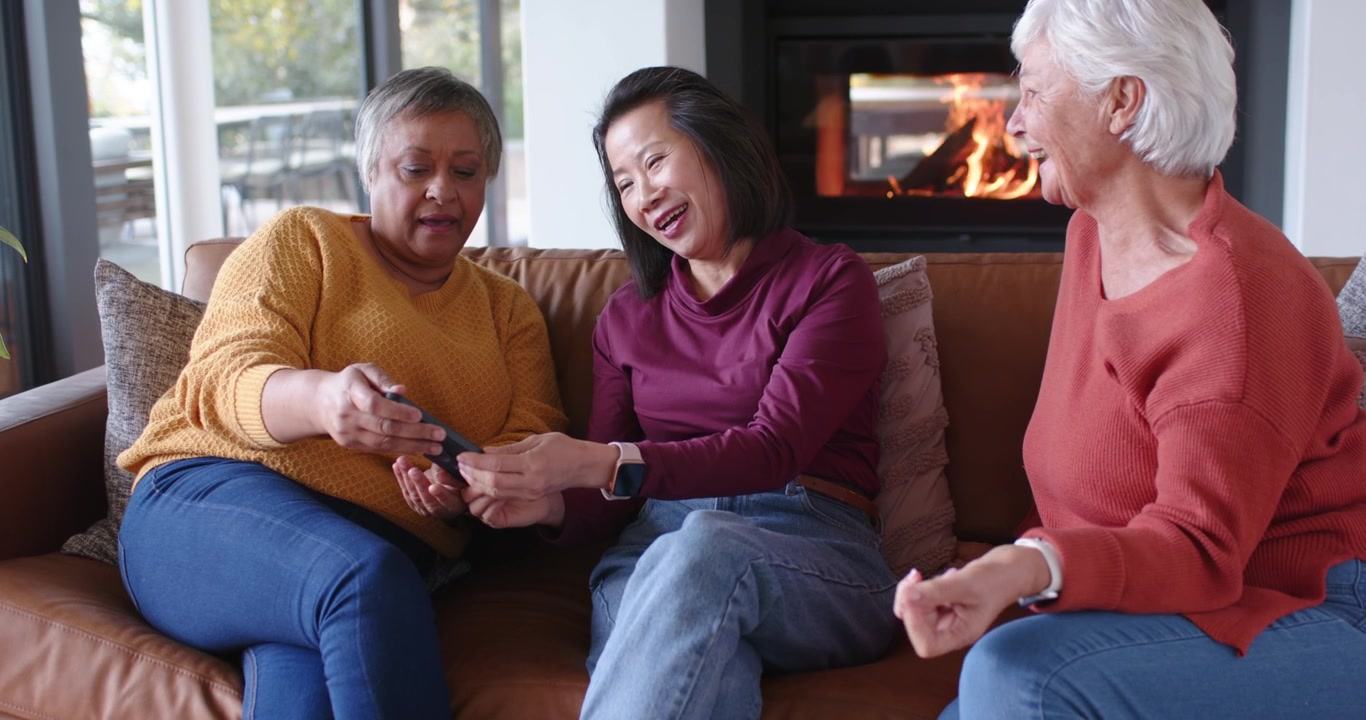 Diverse women friends passing, tapping phone on couch as left woman tilting phone, viewing content