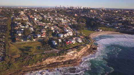 Vista-Aérea-De-La-Playa-De-Bronte-Y-Las-Propiedades-Del-Vecindario-De-Waterfont-Con-El-Horizonte-De-Sydney-Cbd-En-El-Horizonte