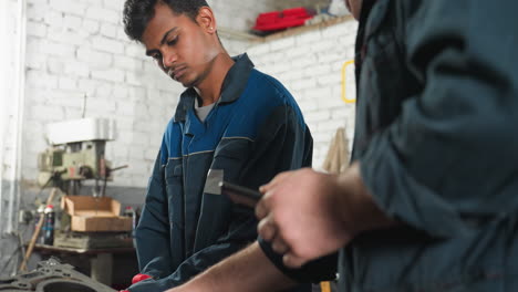 two mechanics in an automotive shop, one working on an engine with an earring, the other showing something on a phone, white car in background