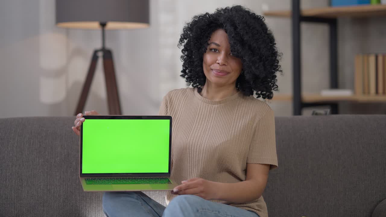 Smiling charming African American woman holding green screen chromakey laptop looking at camera sitting in home office. Portrait of confident positive lady showing startup website sitting on couch.