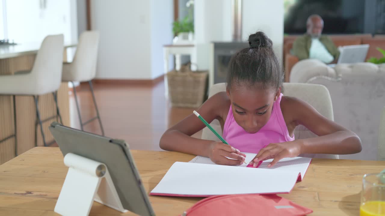 African father and daughter, girl sitting at table drawing in notebook using pink ruler and tablet