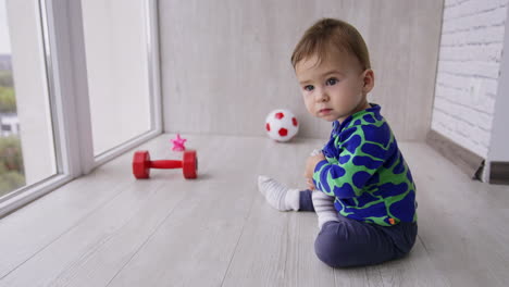 nice cute toddler boy playing on the floor near the big windows. kid wants to take off his sock but then crawls somewhere. toys at backdrop in blur.