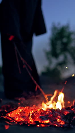 scary witch evil with a stick near the fire. dark death next to smoldered logs of fire in the evening. horror atmosphere outdoors. vertical video