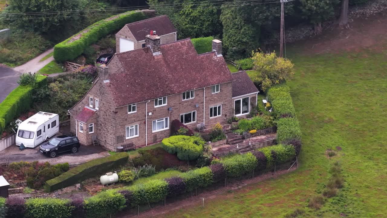 Drone camera smoothly circles a traditional stone farmhouse surrounded by hedges, gardens, and fields in rural England under soft daylight