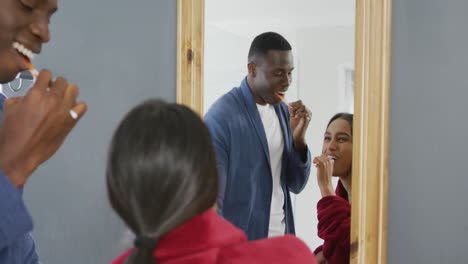 Video-of-happy-diverse-couple-brushing-teeth-looking-in-mirror-and-laughing-in-bathroom-at-home