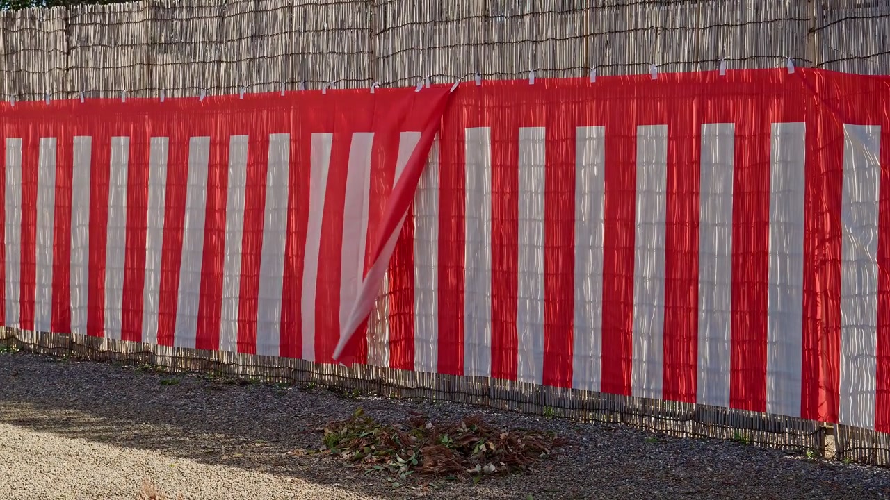 Ein langer rot-weiß gestreifter Kohaku-Vorhang erstreckt sich entlang eines Bambuszauns an einem Shinto-Schrein, dessen Stoff sanft das Licht einfängt und ein traditionelles japanisches Fest oder eine zeremonielle Veranstaltung ankündigt.