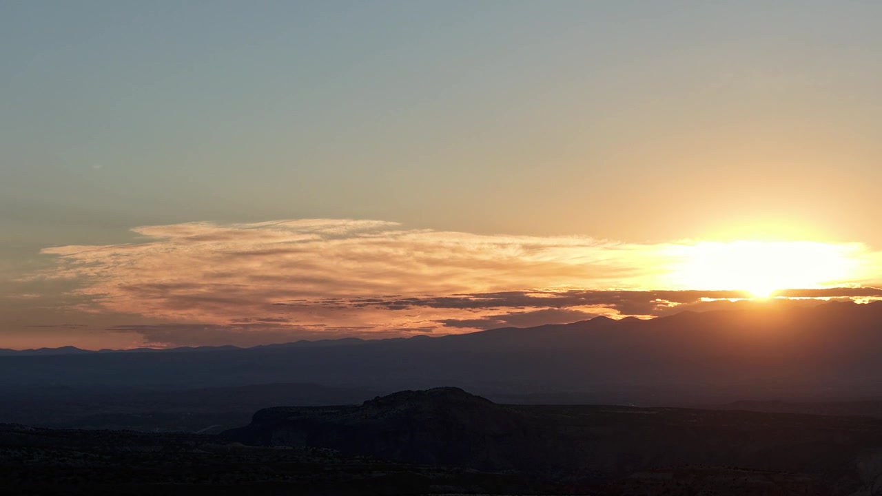 Timelapse aéreo en 4K de un amanecer espectacular en el Suroeste americano. El brillante sol matutino asoma sobre una cordillera distante, proyectando un resplandor dorado y silueteando un primer plano profundo y accidentado.