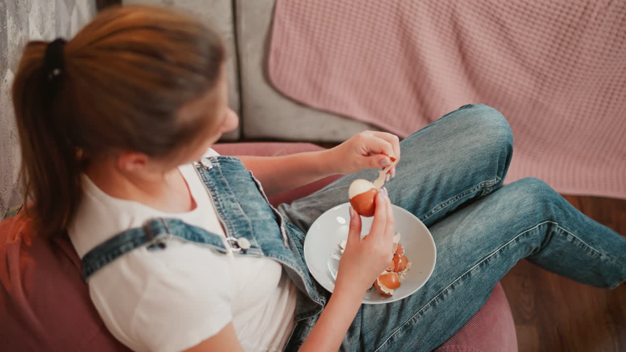 Aerial view of girl seated on pink bean bag peeling brown egg into white plate on lap, surrounded by cozy furnishings and soft indoor lighting in casual relaxed home setting