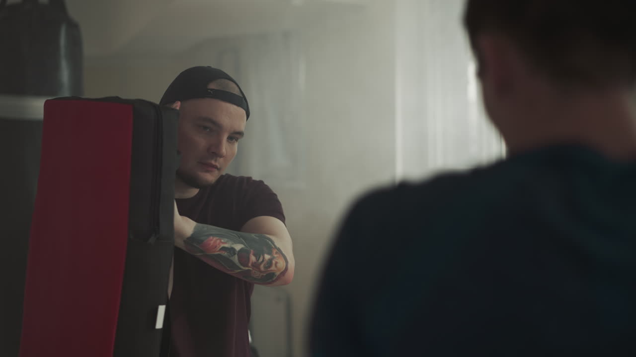 스포츠 클럽의 지하실에서 운동 선수가 맨발로 목표물을 격렬하게 공격합니다. 문신 장비를 가진 남성 강사는 희미하게 조명된 장소에서 학생을 성공으로 안내합니다.