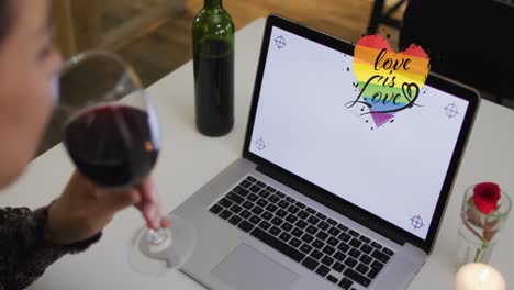 Animation-of-love-is-love-text-over-biracial-woman-drinking-wine-and-using-laptop-with-copy-space