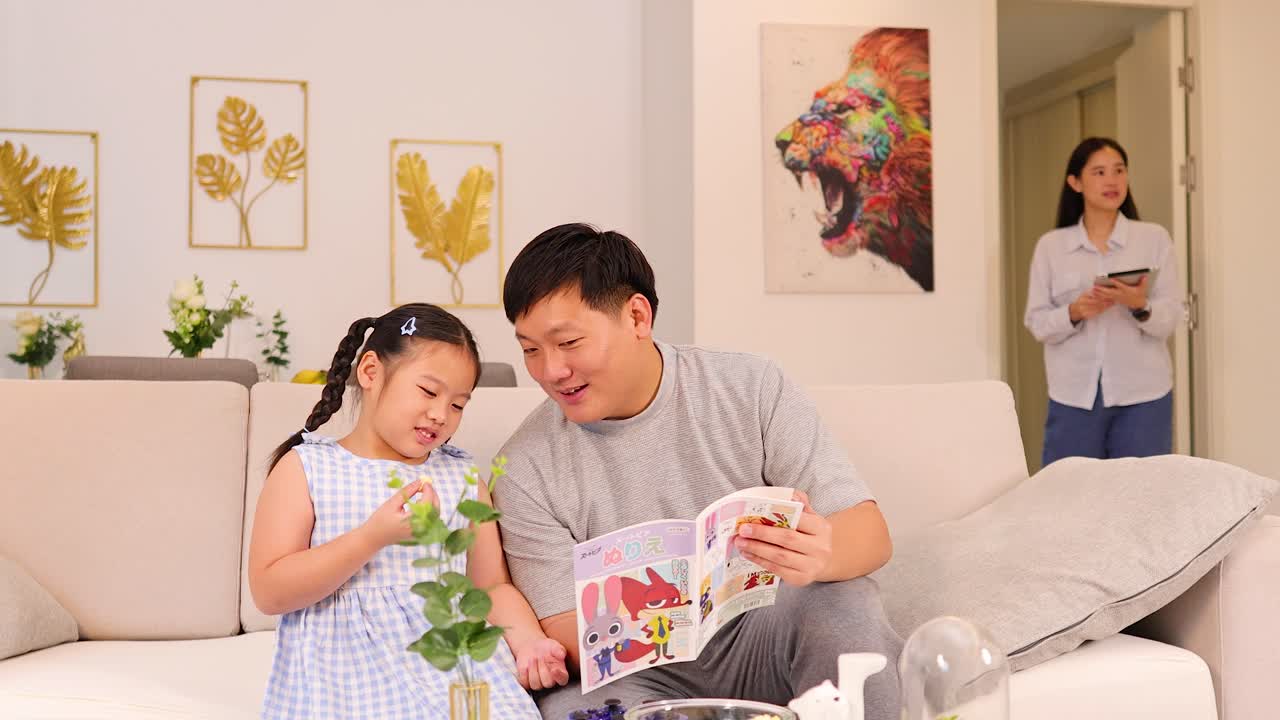 Father and daughter read picture book on couch; mother enters, warm natural light, cozy home