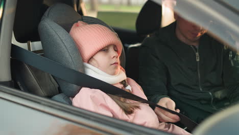 a man in a green jacket is seen carefully buckling the seat belt of his young daughter, who is dressed in a pink jacket and a matching pink beanie, as she sits calmly in a car