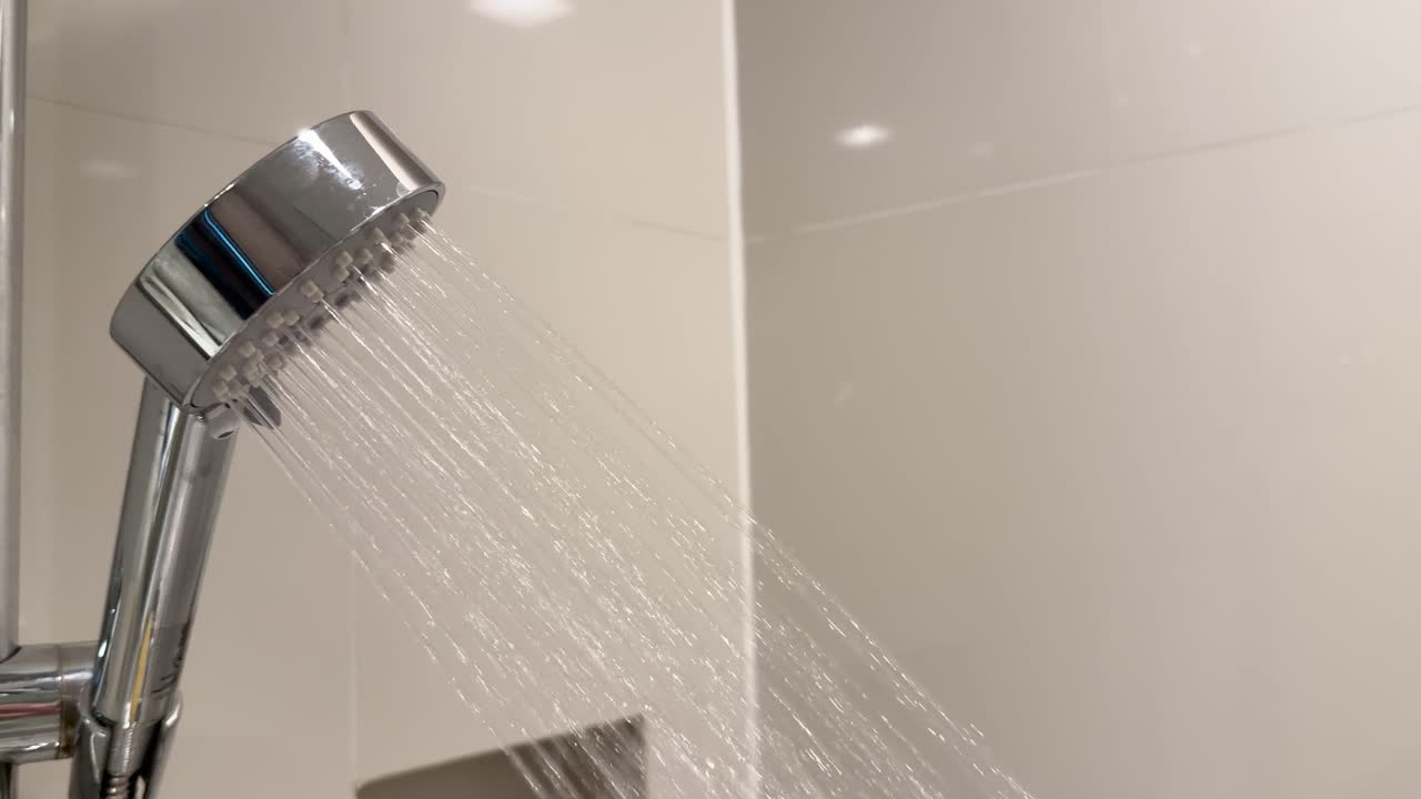 Handheld shower head spraying water in slow motion, bright bathroom lighting, tiled wall background