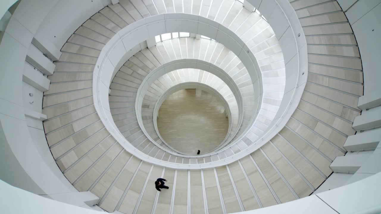 Uomo con cappotto scuro che cammina, entra dall'alto a sinistra e scende una rampa a spirale verso l'atrio centrale con ringhiere bianche.