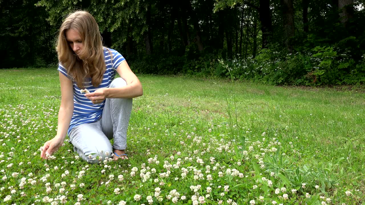 금발의 여자가 풀에 앉아 꽃에서 머리 왕관을 는다. 정적.