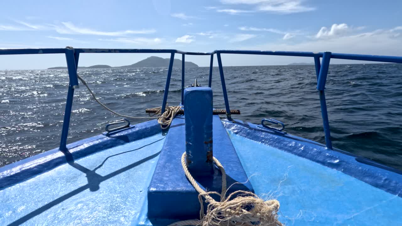Blue boat bow moves through sunlit ocean waves toward distant island, handheld camera, midday lighting