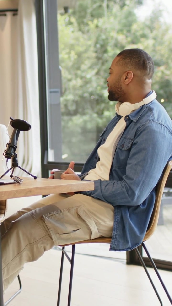 Vertical video: Speaking man in denim shirt gesturing after off-frame podcast prompt at studio, mic