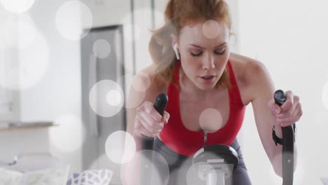 Animation-of-white-spots-over-caucasian-woman-exercising,-using-stationary-bike-at-home