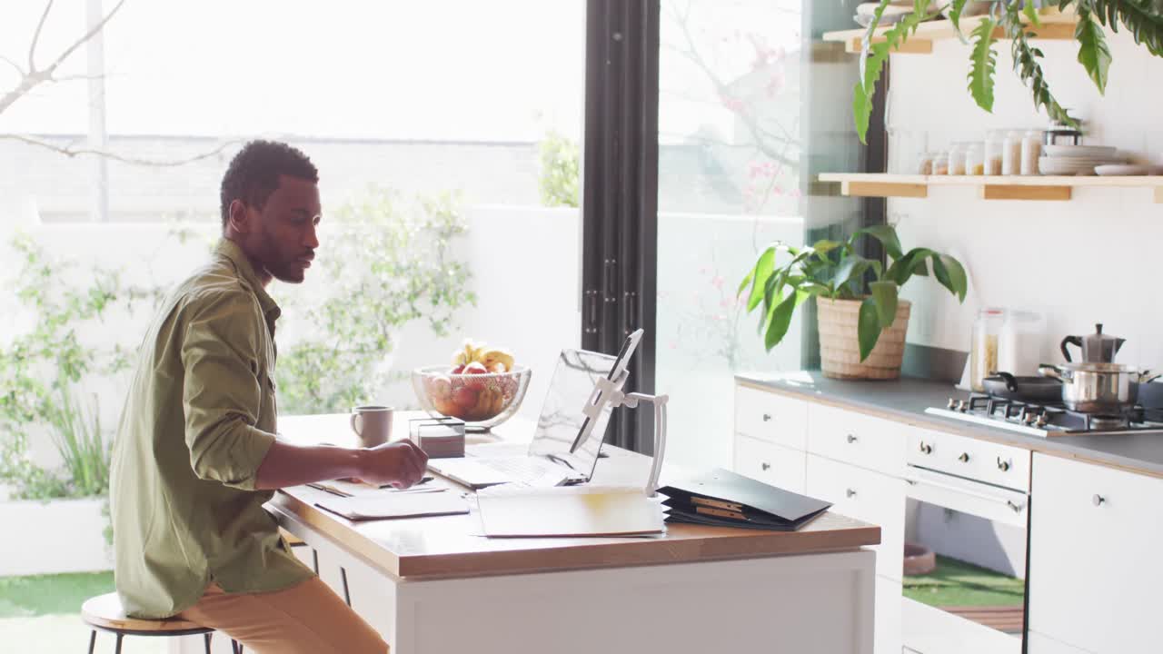 hombre afroamericano sentado en la mesa en la cocina, usando una computadora portátil y tomando notas