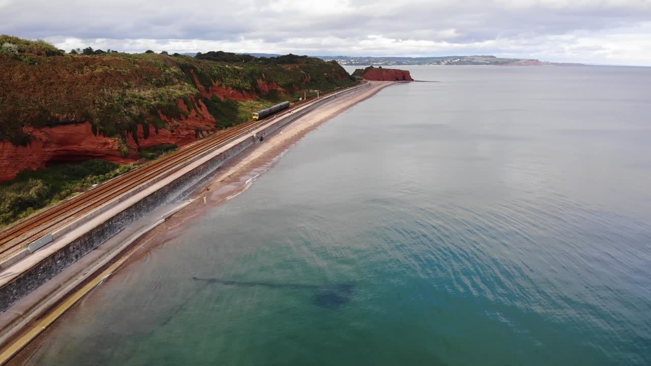 Aerial view of a train passing along the coast in Dawlish, a seaside town in Devon, UK, showing the railway line built on the sea wall. push forward, panning shot