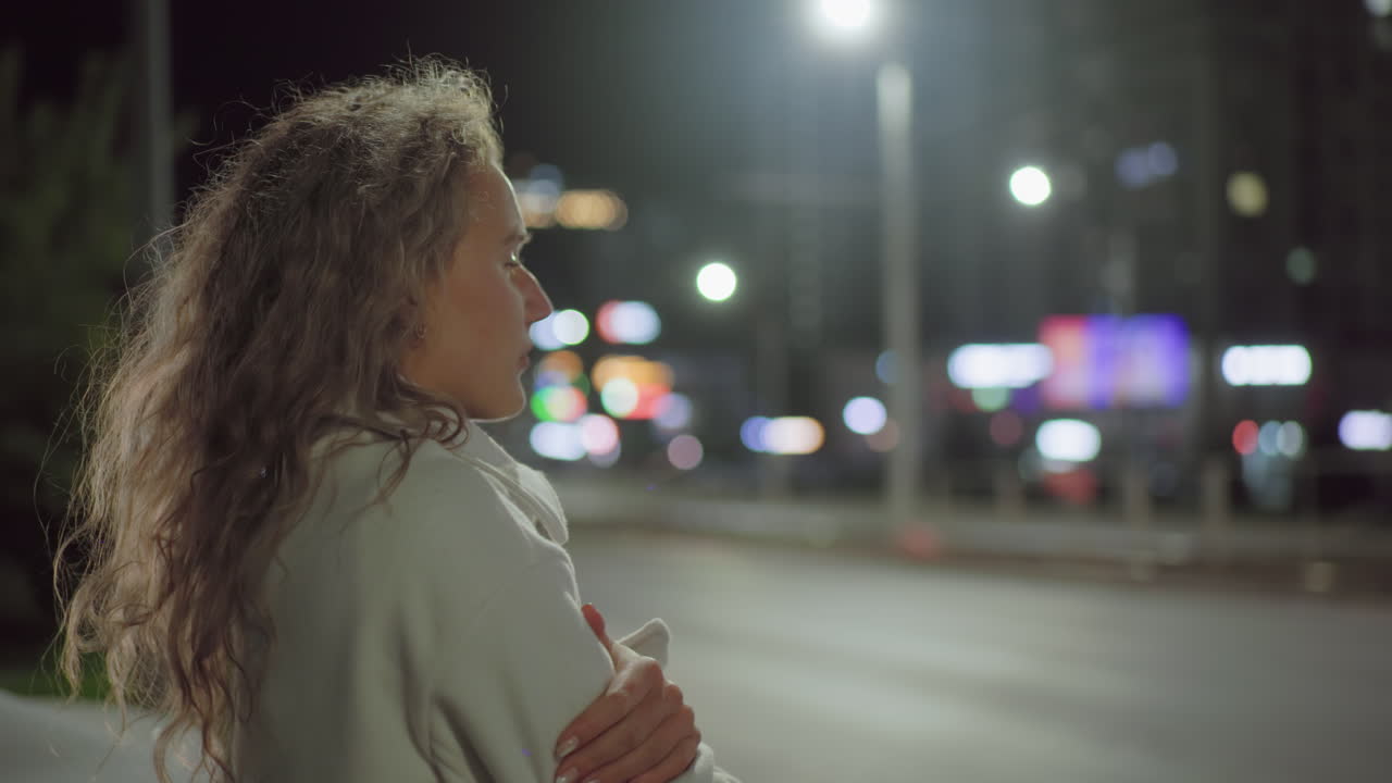 Side view of woman in white coat standing by roadside during cold night with vehicle passing by, illuminated by streetlights and surrounded by blurred urban lights and office buildings