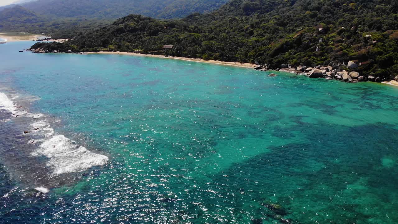 Aerial view overlooking shallow, blue water, rocks and beaches on the coast of Tayrona national natural park, sunny evening, in the Caribbean region of Colombia - tracking, drone shot