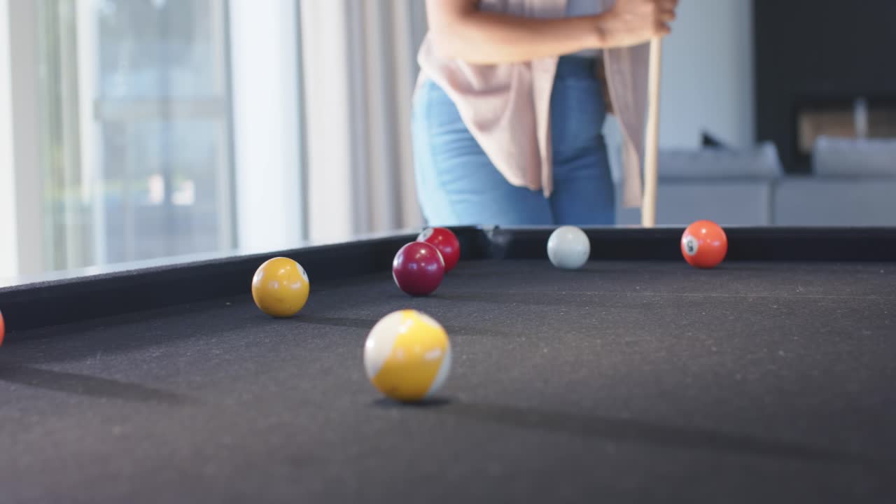 Amiche multietniche che giocano a biliardo in una sala lounge, che mirano i tiri, la stecca che colpisce la bilia bianca e manda in buca le palle.