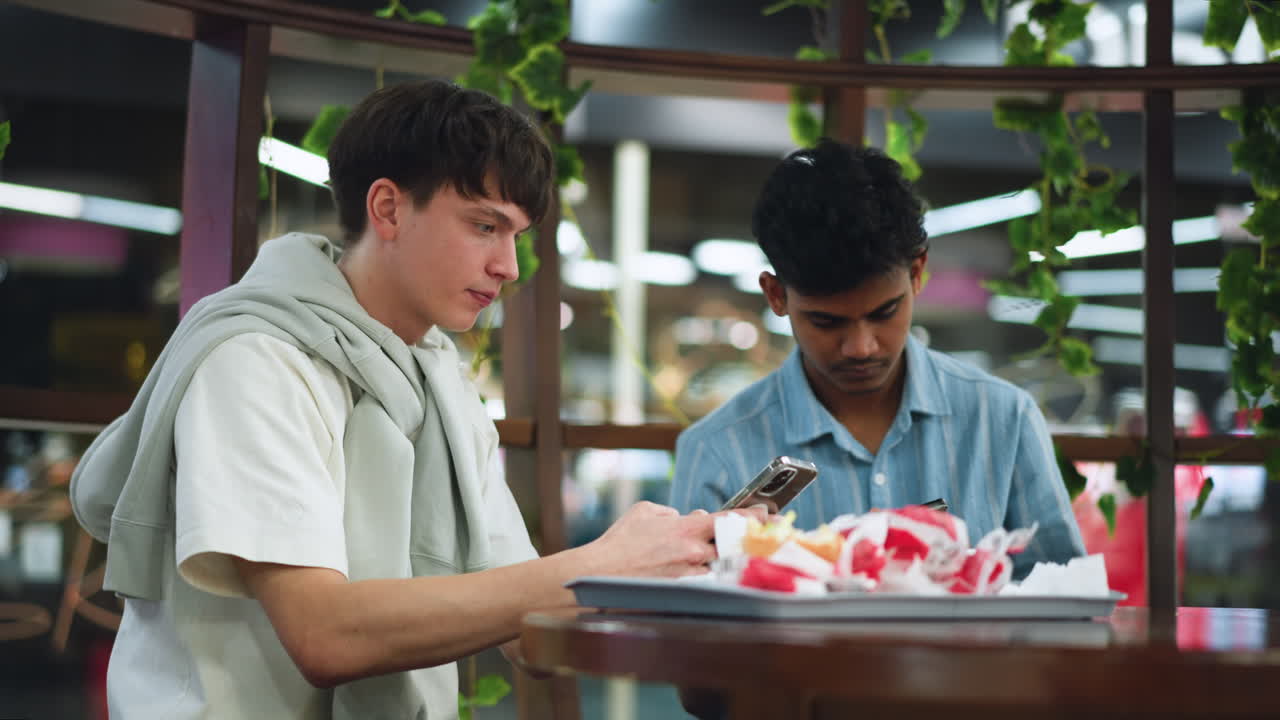View of table with snack bag while two young adults pressing phone together sharing video content and having close interaction in casual indoor setting, focus on phone screen and food