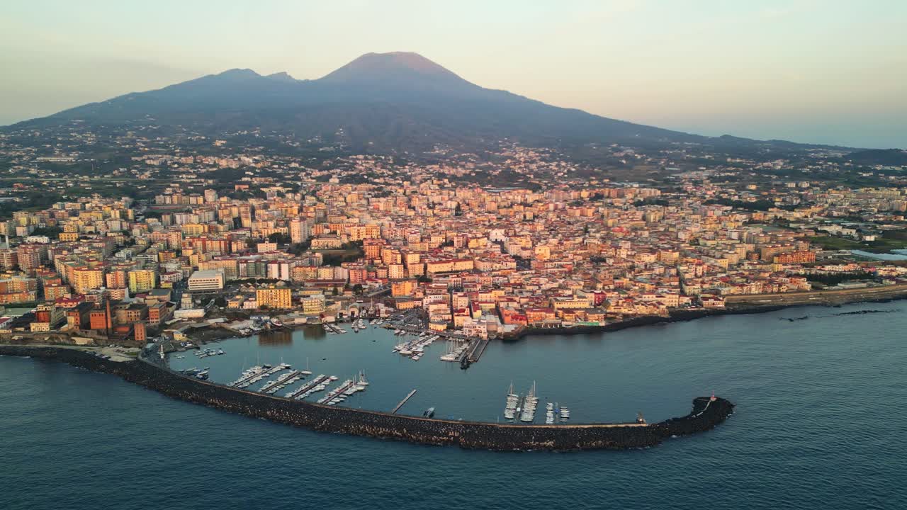 A breathtaking drone shot showing an Italian harbour town at sunrise, with sunlit coastal houses, a peaceful harbour and majestic mountains in the backdrop highlighting the beauty of the Mediterranean