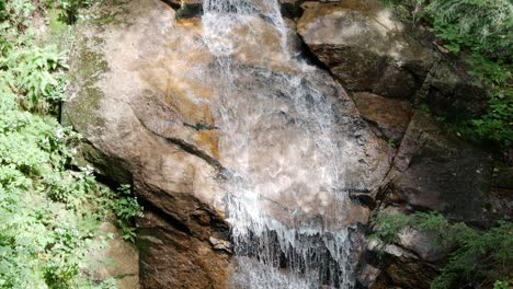 El-Agua-Clara-Fluye-Por-Las-Rocas-Y-Crea-Una-Pequeña-Cascada-Entre-Un-Bosque,-Mientras-La-Cámara-Se-Inclina