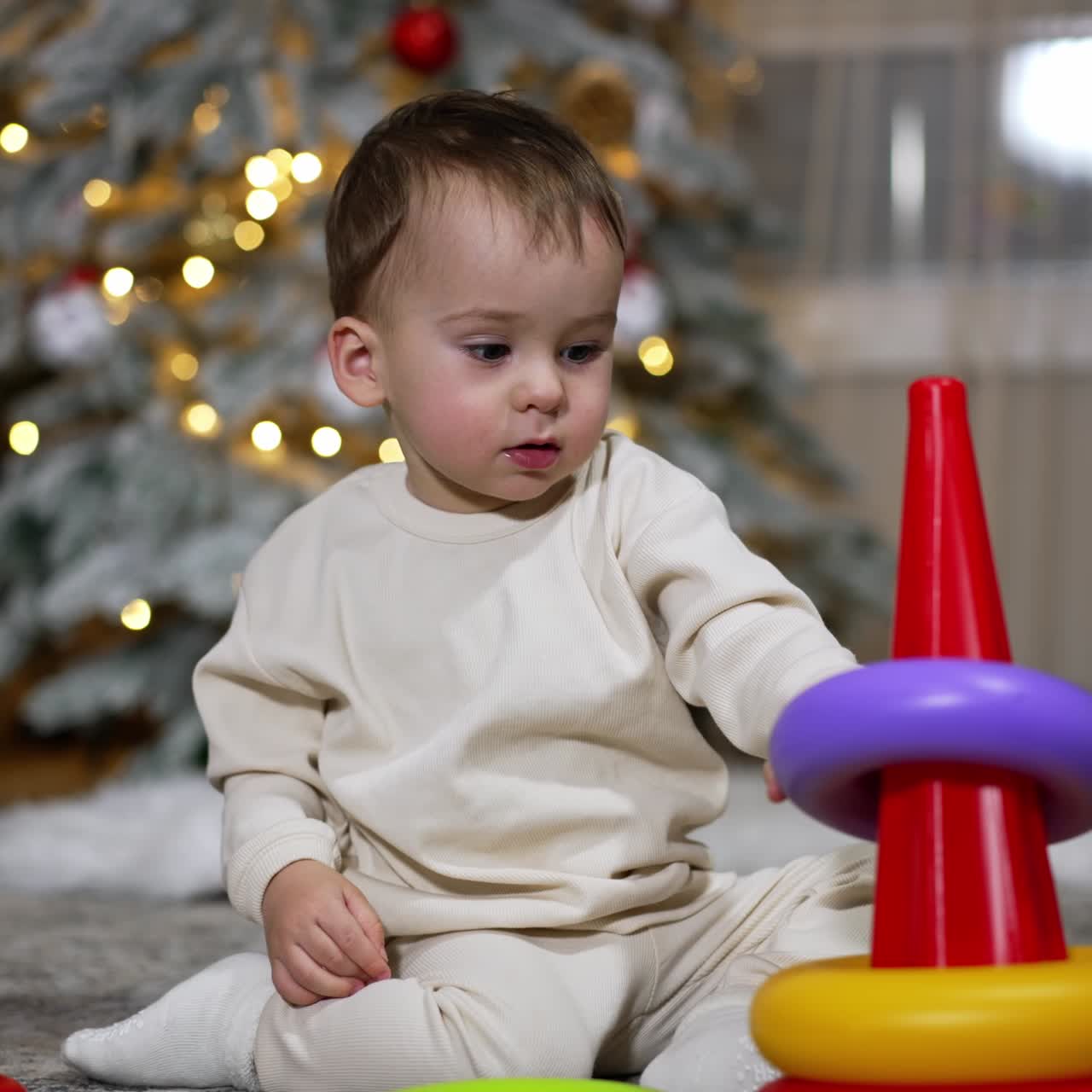 Nice calm baby boy plays with a toy pyramid sitting on the floor. Room decorated for Christmas at backdrop