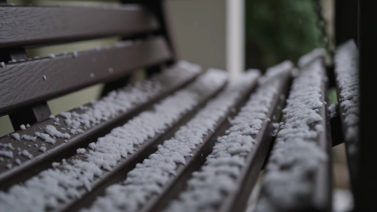 Storm producing sleet pellets falling and gathering on wooden slatted bench on porch near rail post